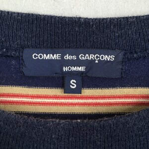 Puede incluir: Primer plano de una camiseta azul marino y a rayas. La camiseta presenta una etiqueta con el texto "COMME des GARCONS HOMME" y la talla "S". La camiseta tiene rayas horizontales en beige y rojo.
