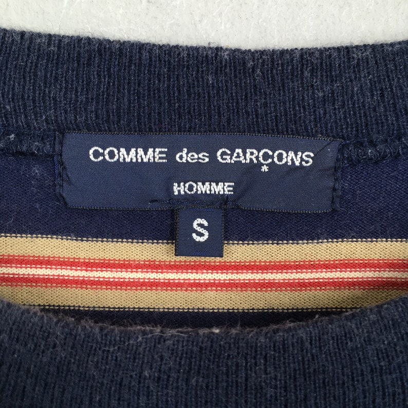 Puede incluir: Primer plano de una camiseta azul marino y a rayas. La camiseta presenta una etiqueta con el texto "COMME des GARCONS HOMME" y la talla "S". La camiseta tiene rayas horizontales en beige y rojo.