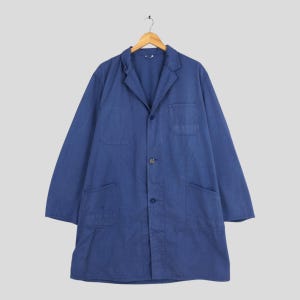 May include: A long, blue work coat with a notched collar, three buttons, and two large front pockets. The coat is hanging on a wooden hanger against a light gray background. The coat's fabric appears to be a durable cotton or linen blend.