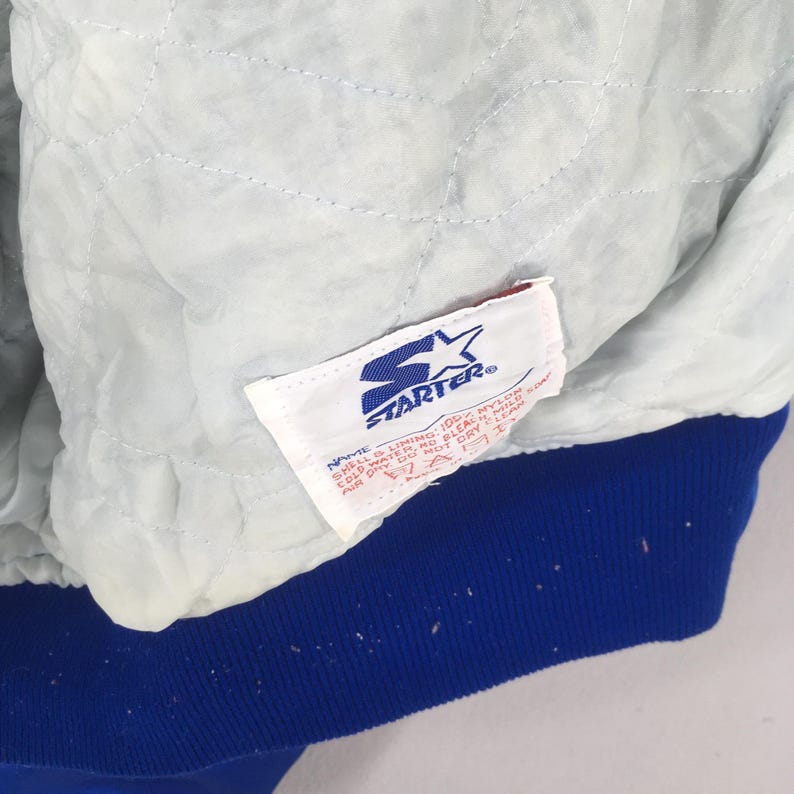 May include: The interior of a jacket featuring a light grey quilted lining and a white Starter brand label. The label displays the blue Starter logo and text including "100% NYLON" and care instructions. A solid blue ribbed hem is visible at the bottom.
