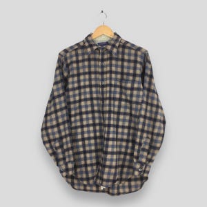 K&ouml;nnte beinhalten: Ein lang&auml;rmeliges Button-Down-Hemd mit einem Karomuster in Blau-, Beige- und Schwarzt&ouml;nen. Das Hemd h&auml;ngt an einem Holzb&uuml;gel vor einem neutralen Hintergrund. Der Kragen und die Manschetten sind sichtbar.