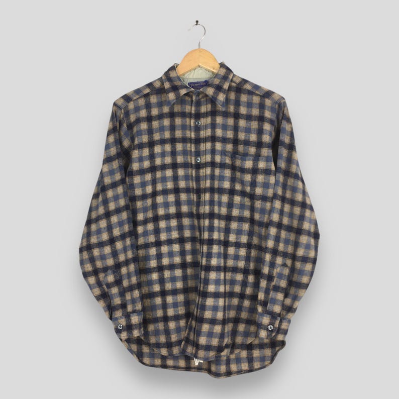K&ouml;nnte beinhalten: Ein lang&auml;rmeliges Button-Down-Hemd mit einem Karomuster in Blau-, Beige- und Schwarzt&ouml;nen. Das Hemd h&auml;ngt an einem Holzb&uuml;gel vor einem neutralen Hintergrund. Der Kragen und die Manschetten sind sichtbar.