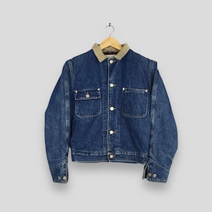 May include: A blue denim jacket with a tan corduroy collar hangs from a wooden hanger. The jacket has two chest pockets, two lower pockets, and button closures. The sleeves have buttoned cuffs.