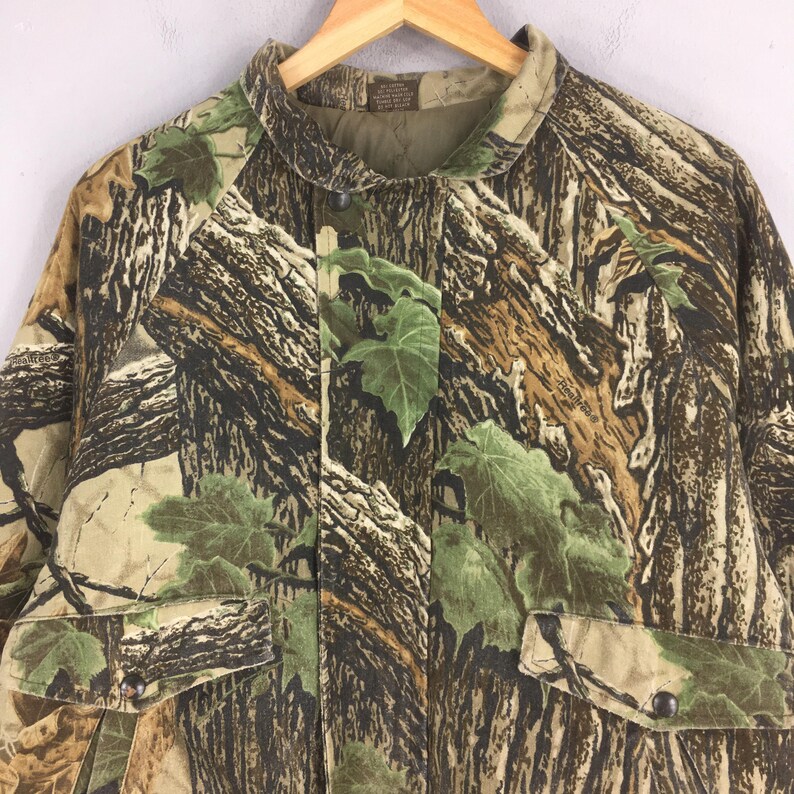 May include: Camouflage jacket with a button-down front, two front pockets, and a collared neckline. The jacket features a realistic tree and leaf pattern in shades of brown, green, and tan. The jacket is hanging on a wooden hanger.