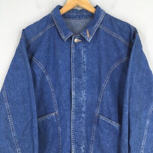 May include: A blue denim jacket with a button-up front, two front pockets, and a classic collar. The jacket is hanging on a wooden hanger. The stitching is visible, and the denim has a slightly faded appearance.