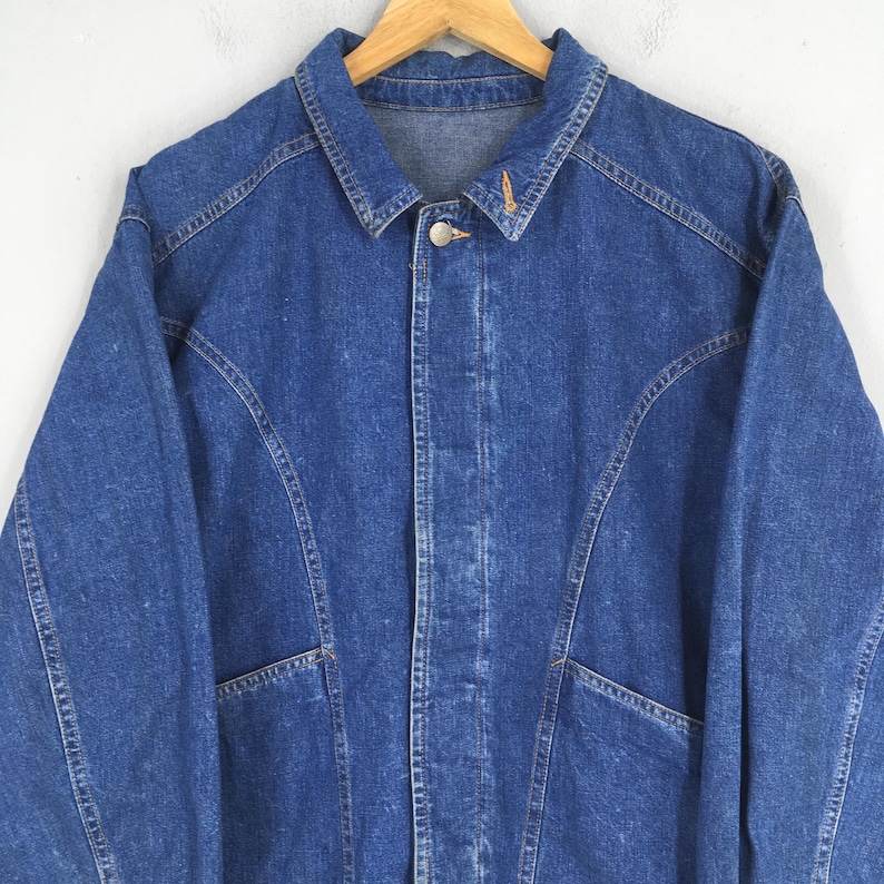 May include: A blue denim jacket with a button-up front, two front pockets, and a classic collar. The jacket is hanging on a wooden hanger. The stitching is visible, and the denim has a slightly faded appearance.
