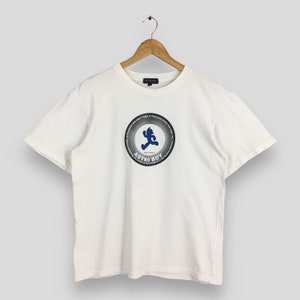 Op de afbeelding: Wit T-shirt met een ronde hals en korte mouwen. Het heeft een cirkelvormige afbeelding van Astro Boy in blauw, met tekst eromheen. Het T-shirt hangt aan een houten hanger.