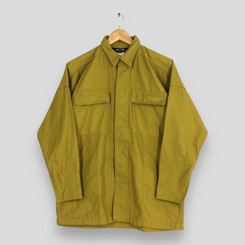 Pode incluir: Camisa verde-oliva de manga comprida com dois bolsos no peito. A camisa &eacute; abotoada com gola e pendurada em um cabide de madeira. A camisa &eacute; feita de um tecido dur&aacute;vel.