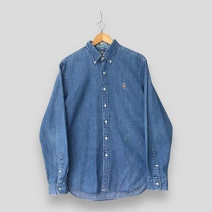 May include: A long-sleeve button-down shirt in a medium blue denim fabric. The shirt features a button-down collar, a front button placket, and a small embroidered logo on the left chest. The cuffs are adjustable with buttons on the sleeves.