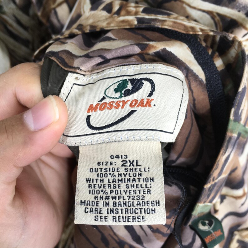 Puede incluir: Primer plano de una etiqueta de chaqueta de camuflaje Mossy Oak. La etiqueta dice "MOSSY OAK" con un logotipo. La talla es 2XL. La chaqueta est&aacute; hecha de nailon y poli&eacute;ster. La etiqueta tambi&eacute;n incluye instrucciones de cuidado.