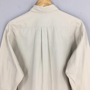 Peut inclure: Chemise boutonn&eacute;e beige clair &agrave; manches longues, vue de dos. La chemise est suspendue &agrave; un cintre en bois. Le col est boutonn&eacute; et il y a un pli au centre du dos.