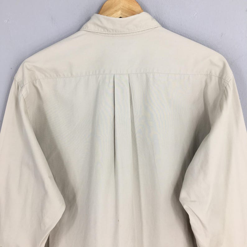 Peut inclure: Chemise boutonn&eacute;e beige clair &agrave; manches longues, vue de dos. La chemise est suspendue &agrave; un cintre en bois. Le col est boutonn&eacute; et il y a un pli au centre du dos.