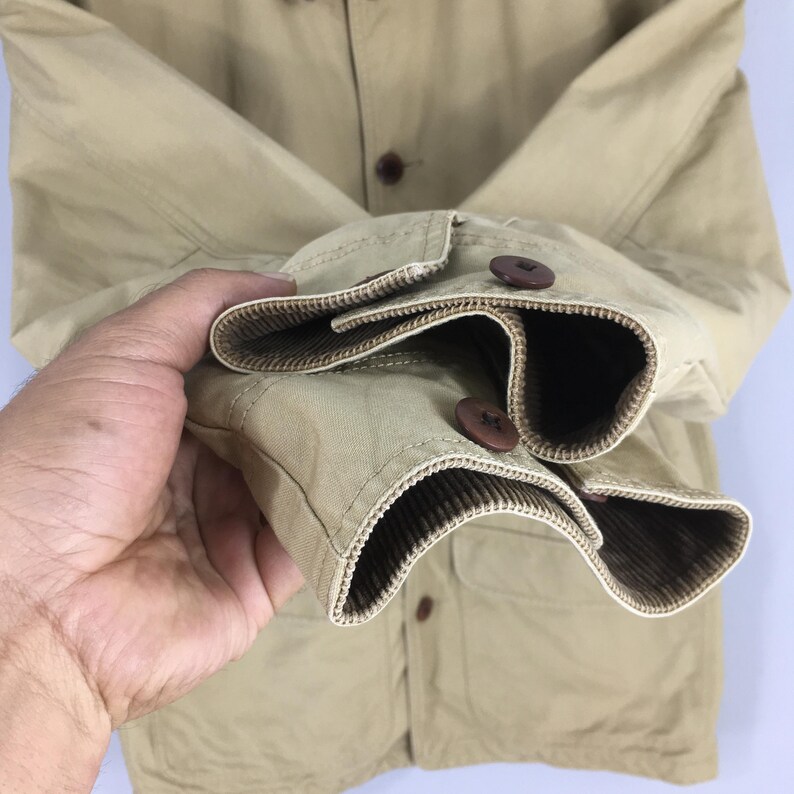 May include: Close-up of a tan jacket with brown buttons and a ribbed cuff detail. The jacket has a classic design with a button-down front and two front pockets. The sleeves are cuffed, revealing a brown ribbed lining.