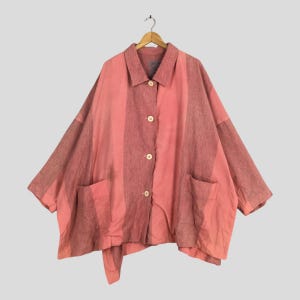 Peut inclure: Une chemise ample et surdimensionnée, dans des tons de rose et de mauve, suspendue à un cintre en bois. La chemise présente un motif à rayures verticales, deux poches avant et des boutons en bois. Le tissu semble être un matériau tissé léger.