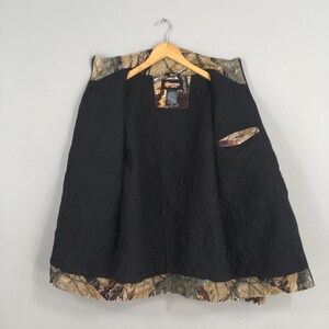 May include: Camouflage pattern jacket with a black quilted lining. The jacket is open, revealing the interior lining and a small pocket. The outer shell features a brown, green, and tan camouflage design. The jacket hangs from a wooden hanger.