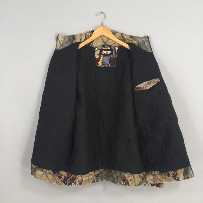 May include: Camouflage pattern jacket with a black quilted lining. The jacket is open, revealing the interior lining and a small pocket. The outer shell features a brown, green, and tan camouflage design. The jacket hangs from a wooden hanger.
