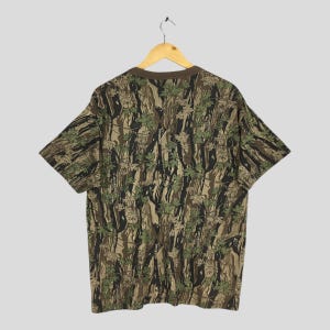 May include: A short-sleeved t-shirt with a camouflage pattern featuring brown, green, and black tones. The shirt is hanging on a wooden hanger against a light gray background. The pattern includes leaf and tree trunk designs.