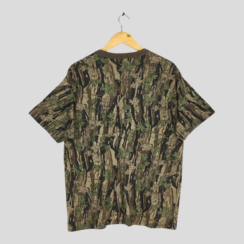 May include: A short-sleeved t-shirt with a camouflage pattern featuring brown, green, and black tones. The shirt is hanging on a wooden hanger against a light gray background. The pattern includes leaf and tree trunk designs.