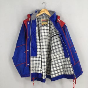 May include: Blue and red vintage FILA Italia Sport jacket with a plaid lining. The jacket has a hood, a zipper, and snap closures. The FILA logo is visible on the chest. The jacket is hanging on a wooden hanger.