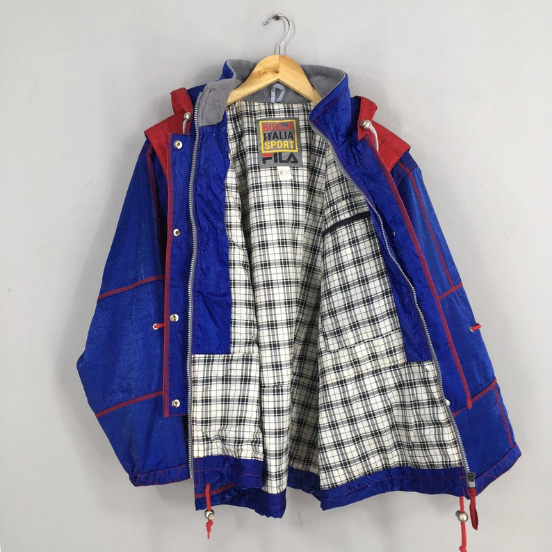 May include: Blue and red vintage FILA Italia Sport jacket with a plaid lining. The jacket has a hood, a zipper, and snap closures. The FILA logo is visible on the chest. The jacket is hanging on a wooden hanger.