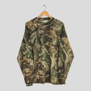 Könnte beinhalten: Langärmeliges Camouflage-Shirt mit Stehkragen. Das Shirt hat ein Muster aus braunen, grünen und beigen Blättern und Ästen. Das Shirt hängt an einem Holzbügel.