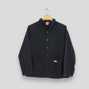 Puede incluir: Chaqueta de trabajo negra con cierre de botones, dos bolsillos delanteros y una pequeña etiqueta roja y blanca. La chaqueta está colgada de una percha de madera sobre un fondo gris claro. La chaqueta tiene cuello.