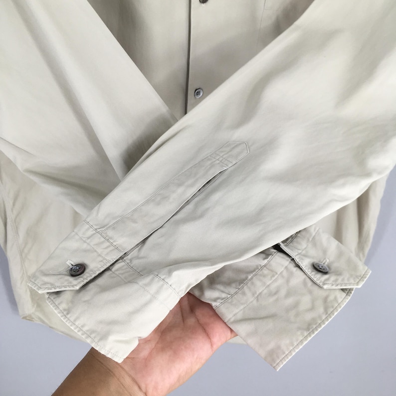 Peut inclure: Gros plan d'une chemise boutonn&eacute;e beige clair. La chemise a des manches longues avec des poignets boutonn&eacute;s et une petite poche sur la manche gauche. Le tissu semble &ecirc;tre l&eacute;ger.