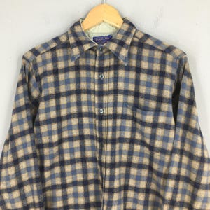 K&ouml;nnte beinhalten: Ein kariertes Flanellhemd in Blau, Beige und Marine. Das Hemd hat einen Kragen, eine Brusttasche und lange &Auml;rmel. Das Pendleton-Etikett ist am Kragen sichtbar.