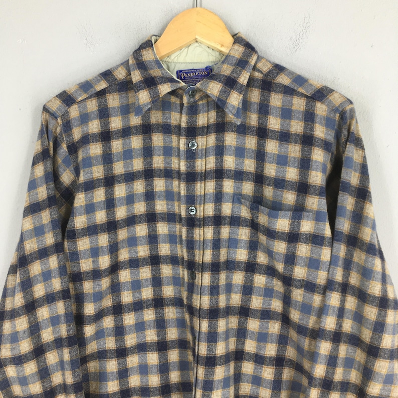 K&ouml;nnte beinhalten: Ein kariertes Flanellhemd in Blau, Beige und Marine. Das Hemd hat einen Kragen, eine Brusttasche und lange &Auml;rmel. Das Pendleton-Etikett ist am Kragen sichtbar.
