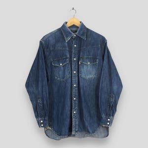 Puede incluir: Una camisa vaquera azul medio con mangas largas, cierre de botones y dos bolsillos en el pecho. La camisa tiene botones blancos y un cuello clásico. La prenda está colgada de una percha de madera.