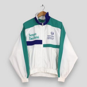 May include: White, green, and navy blue Sergio Tacchini zip-up jacket. The jacket features the brand's logo and name on the front. The jacket has a stand-up collar and ribbed cuffs and hem. The jacket is hanging on a wooden hanger.