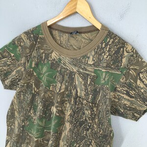 Puede incluir: Una camiseta de camuflaje de manga corta con cuello marr&oacute;n y bolsillo. La camiseta presenta un estampado de hojas verdes y corteza de &aacute;rbol marr&oacute;n, creando un dise&ntilde;o de camuflaje de bosque. La camiseta est&aacute; colgada de una percha de madera.