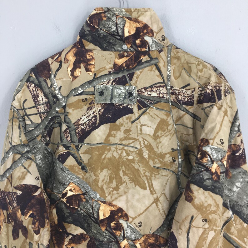 May include: A camouflage jacket in shades of brown, tan, and green, featuring a button-up collar and a small rectangular patch with two buttons. The jacket has a classic outdoor design.