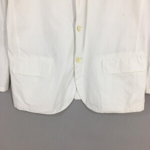 May include: A crisp white blazer featuring a two-button front closure with light-colored buttons. The garment includes two rectangular flap pockets positioned on the lower front panels and a classic notched lapel collar design.