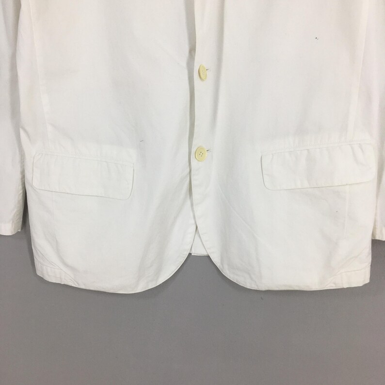 May include: A crisp white blazer featuring a two-button front closure with light-colored buttons. The garment includes two rectangular flap pockets positioned on the lower front panels and a classic notched lapel collar design.