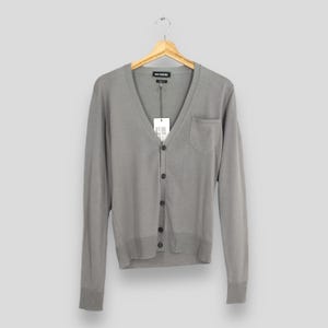 Peut inclure: Un cardigan gris avec un col en V, des manches longues et une seule poche sur le côté gauche. Le pull boutonné est suspendu à un cintre en bois. Une étiquette est attachée au pull.
