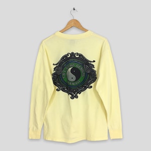 May include: Pale yellow long-sleeve shirt with a large graphic on the back. The graphic features a yin-yang symbol in a green and blue circle, framed by a decorative border. The text "T&C Surf Designs Hawaii" is also visible.