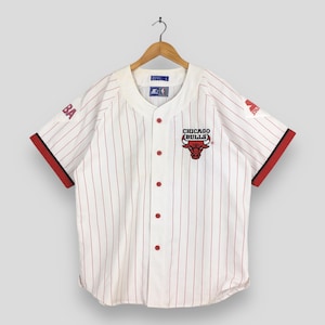 Puede incluir: Camiseta de béisbol blanca con finas rayas rojas, con el logo de los Chicago Bulls en el pecho. La camiseta tiene detalles rojos en las mangas y botones. El logo de Starter es visible en la manga.