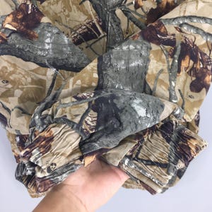 May include: Close-up of a camouflage jacket featuring a pattern of brown, green, and gray hues. The fabric has a textured appearance, with details resembling tree bark and foliage. The jacket's sleeve cuff is visible, showing gathered fabric.