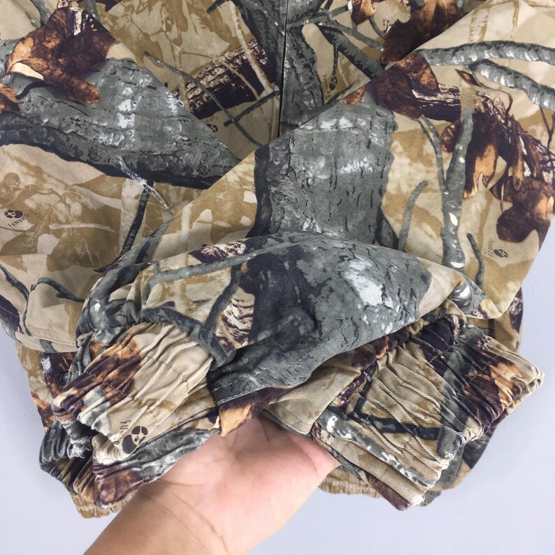 May include: Close-up of a camouflage jacket featuring a pattern of brown, green, and gray hues. The fabric has a textured appearance, with details resembling tree bark and foliage. The jacket's sleeve cuff is visible, showing gathered fabric.