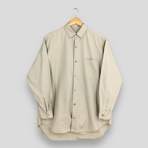 Peut inclure: Une chemise beige boutonn&eacute;e &agrave; manches longues avec une poche poitrine. La chemise est suspendue &agrave; un cintre en bois sur un fond gris clair. La chemise a un col classique et des poignets boutonn&eacute;s.