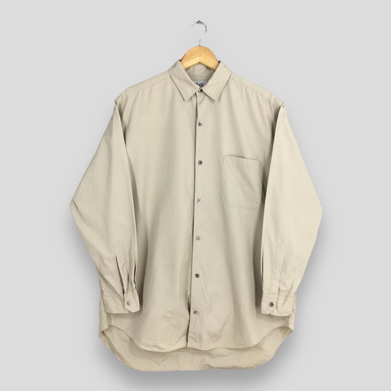 Peut inclure: Une chemise beige boutonn&eacute;e &agrave; manches longues avec une poche poitrine. La chemise est suspendue &agrave; un cintre en bois sur un fond gris clair. La chemise a un col classique et des poignets boutonn&eacute;s.