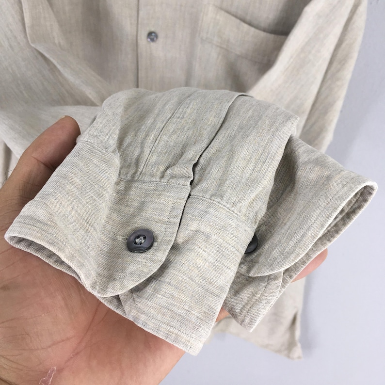 May include: Close-up of a light beige long-sleeved shirt. The shirt features a button-down front, a chest pocket, and buttoned cuffs. The fabric has a textured appearance, and the buttons are a dark gray color.