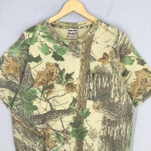 May include: A short-sleeved t-shirt with a camouflage pattern featuring brown, green, and tan colors. The shirt has a pocket on the chest and a crew neck. The tag reads "Outdoors".