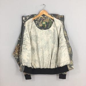 Op de afbeelding: Een camouflage-printjas met een zwarte rits en een zwarte tailleband. De jas heeft een witte voering en twee binnenzakken. Het label op de jas luidt "Cabela's".