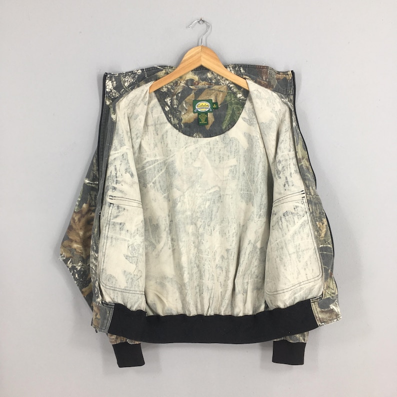 Op de afbeelding: Een camouflage-printjas met een zwarte rits en een zwarte tailleband. De jas heeft een witte voering en twee binnenzakken. Het label op de jas luidt "Cabela's".