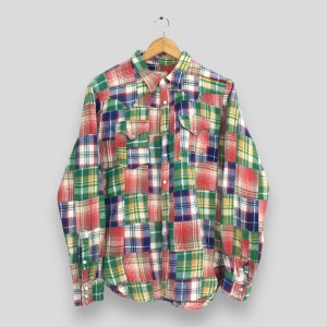 Pode incluir: Uma camisa de manga comprida em patchwork numa variedade de cores, incluindo vermelho, verde, azul e amarelo. A camisa tem uma frente abotoada, dois bolsos no peito e uma gola clássica. A camisa está pendurada num cabide de madeira.