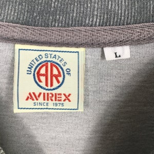 May include: Close-up of a gray t-shirt collar with a woven texture. The shirt features a rectangular label with the text "UNITED STATES OF AVIREX SINCE 1975" in red and blue. A white label with the letter "L" is also visible.