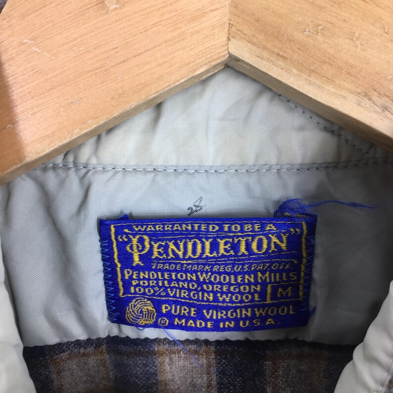 K&ouml;nnte beinhalten: Nahaufnahme eines blauen und goldenen Pendleton-Etiketts. Das Etikett lautet "Warranted to be a Pendleton" und "100% Virgin Wool". Das Etikett ist in ein braun-beiges kariertes Hemd eingen&auml;ht.