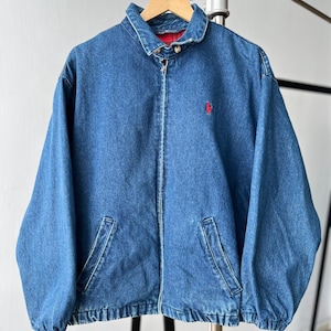 May include: A blue denim jacket featuring a full-length front zipper, a fold-down collar, and two slanted front pockets. The jacket has a red embroidered logo on the left chest and a red and black plaid lining visible at the inner collar area.